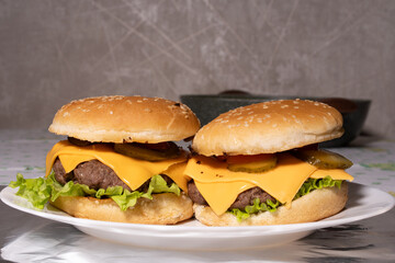 Two cheeseburgers with sesame buns, beef patties, melted cheddar, lettuce, pickles, and tomato sit on a white plate, ready for serving.