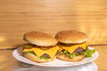Two cheeseburgers with beef patties, cheddar cheese, pickles, lettuce, and tomatoes on sesame buns, served on a white plate with a rustic background.