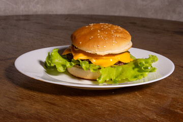 A juicy cheeseburger with a sesame seed bun, melted cheese, beef patty, and lettuce on a white plate, styled atop a rustic wooden background.