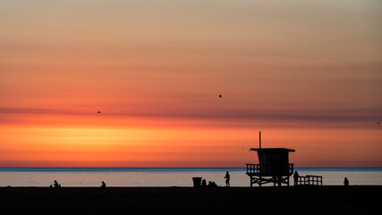 A beautiful sunset casts vibrant colors across a tranquil beach scene, highlighting silhouettes of people and iconic lifeguard station against the calming ocean backdrop.