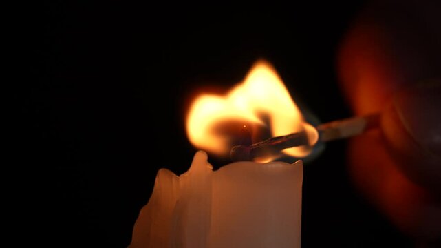 Male hand lights paraffin candle using a match on a black background, close up. White candle with orange fire. Bright flame, fire, ignition in a dark room. Concept of memory, celebration, ritual
