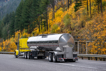 Gorgeous classic big rig yellow semi truck transporting liquid cargo in stainless steel tank semi...