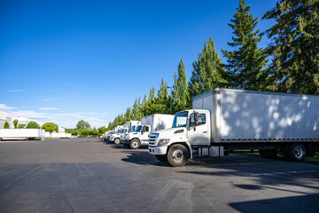 Industrial grade middle duty day cab rigs semi trucks with box trailers standing in row on warehouse parking lot waiting for the next local deliveries