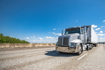 Low profile gray big rig semi truck transporting cargo in refrigerator semi trailer running on the wide multiline highway road with concrete protection barrier