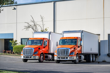Orange day cab middle duty rigs semi trucks with box trailers standing in warehouse dock gates...