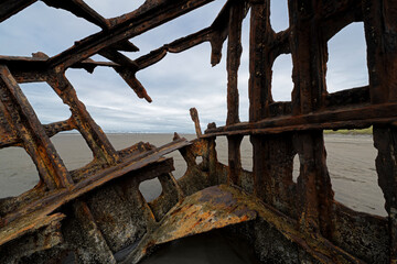 Inside the decaying hull of a shipwreck, the rusty structure tells stories of ocean adventures, surrounded by sandy shores and distant hills under an overcast sky.