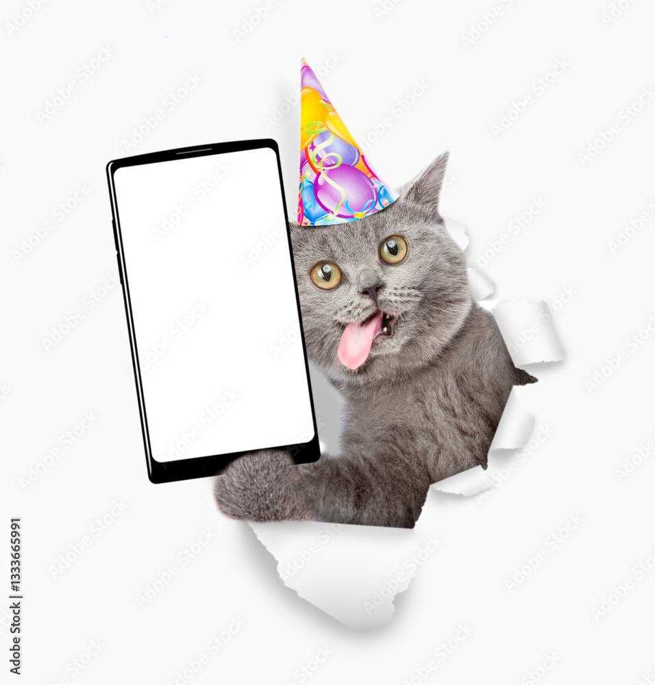 Poster Crazy cat with tongue out wearing party cap showing smartphone with white blank screen in it paw through torn white paper hole. Empty free space for mock up, banner