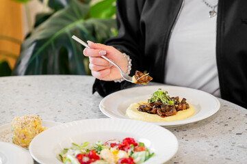 A close-up of a hand holding a fork with a delicious dish of creamy mashed potatoes topped with mushrooms. In the foreground, fresh salads and a side dish are presented on a table.