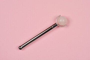 A silver facial roller with a pink quartz ball at the end lies diagonally on a pink background