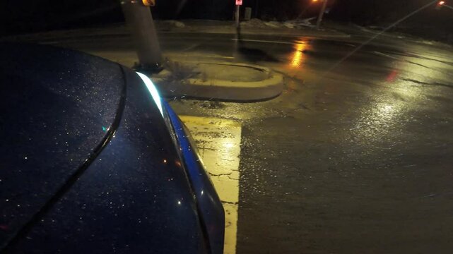 A headlight signaling left on a blue car coming to a stop at a traffic light on a rainy night