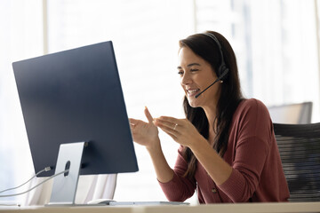 Positive Latin businesswoman in headphones talking on video call at monitor, speaking, laughing, moving hands. Cheerful online teacher giving business training, webinar