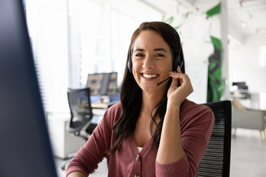 Happy customer contact center employee posing at workplace, touching headset with microphone, looking at camera, smiling. Positive Latin businesswoman, support service operator portrait