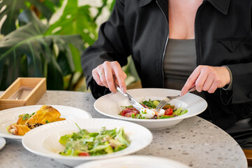 A close-up of a person enjoying a fresh salad at a table, surrounded by various dishes. The focus is on the hands using a fork and knife, showcasing a healthy meal in a vibrant atmosphere.
