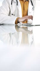 Unknown doctor man in a yellow shirt under a medical coat is using technology in medicine. Close-up of hands typing on a laptop on a glass desk in the clinic, vertical view. Health care concept