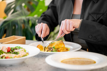 A person enjoys a delicious meal at a stylish restaurant, elegantly cutting into a gourmet dish surrounded by fresh salad and soup. Green plants add a vibrant touch.
