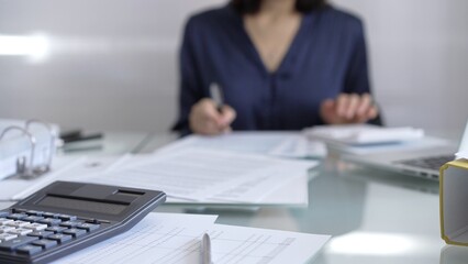Black calculator, pen and documents are lying on accountant's desk. Businesswoman wearing blue dress is working with documents on the background. Business and audit concept
