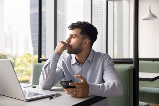 Pensive, thoughtful young male freelancer sit at desk with laptop in modern coworking office holding phone waiting for response, reviewing received information, or considering work or personal matter