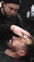 Fototapeta premium Professional barber carefully shaving client's beard with straight razor, applying gel and using hot towel for precise grooming in contemporary barbershop setting, vertical view