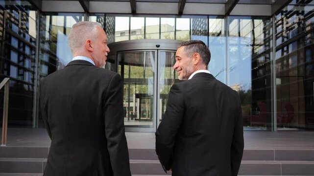 Two well-dressed businessmen stroll confidently, talking in a lively discussion as they walk towards a corporate office entrance, showcasing their professionalism in the office workplace
