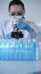 Female scientist wearing lab coat and protective blue gloves using microscope doing research in...