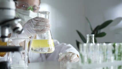 Woman scientist wearing a lab coat, white gloves, and a mask is pouring a yellow oily liquid from one beaker to another in a laboratory setting, close up. Science and medicine