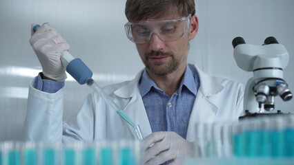 Scientist man wearing a lab coat and safety glasses is using a micropipette to drop a liquid into a...