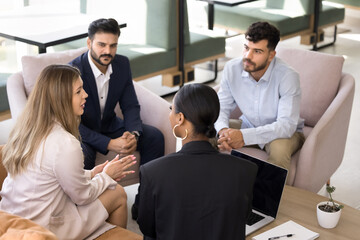 Overhead top view four young and ambitious professional team negotiate seated in modern office lobby, listening young European teammate presenting new project details, develop start up ideas. Teamwork