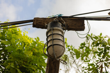 A vintage industrial outdoor lamp with protective metal cage, mounted on a weathered wooden post with electrical wires. Lush green foliage in the background.