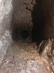 Inside a dim rocky tunnel, rough stone walls slope inward and lead to a circular opening. The...