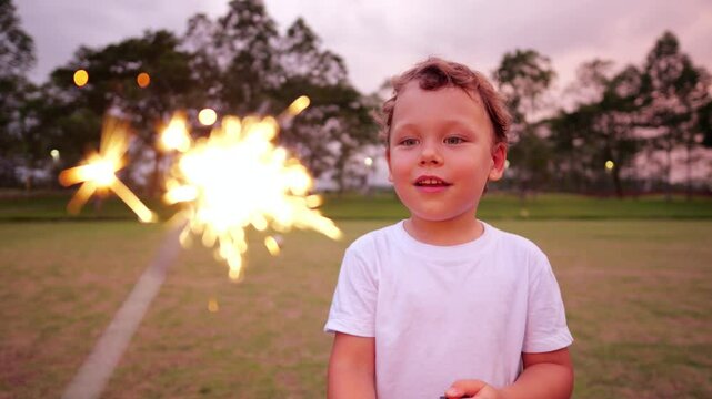 Young boy glowing with excitement and fascinated by sparkler stick, which is glowing near camera, seen close in blur. Little one experiencing magical childhood moment during festive evening