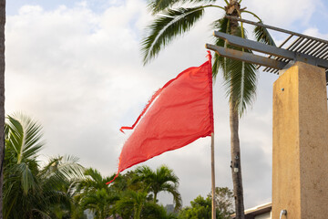 A tattered red warning flag waving in the wind, set against palm trees and a partly cloudy sky. Cautionary symbol for danger, high winds, or hazardous conditions