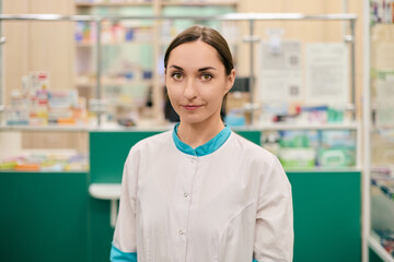 Young female pharmacist with dark hair and caucasian ethnicity stands confidently. Pharmacist wears white coat, surrounded by shelves of medicines in well-lit pharmacy
