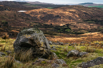 A large rock sits in the foreground, surrounded by grass and scattered stones, while rolling hills...