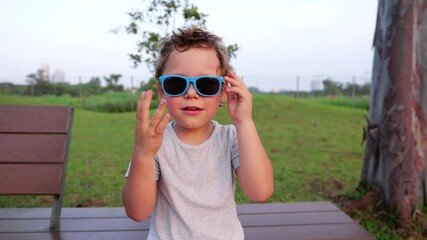 Cheerful little boy wearing colorful plastic sunglasses while sitting on park bench, making playful expression and enjoying carefree moment on summer evening. Portrait shot of funny little kid