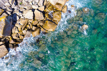 Top-down image of a rocky shore meeting the sea, stunning green waters and summer vacation vibes background