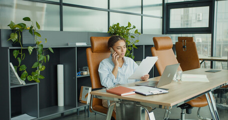 A young professional woman multitasking in a modern office, talking on the phone while analyzing documents at a desk with a laptop, plants, and stylish furniture