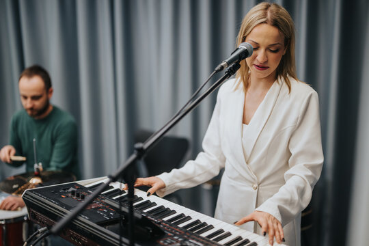 A female musician performs on the keyboard while singing into a microphone, accompanied by a drummer during a band practice session in a professional studio setting.