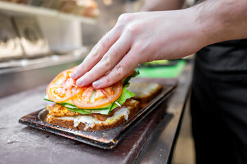 A hand carefully places fresh tomato slices on a sandwich filled with vegetables and melted cheese, highlighting the art of food preparation in a bustling kitchen setting.