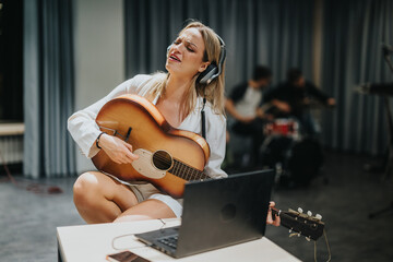 A woman in a music studio wears headphones while playing the guitar. She passionately practices...