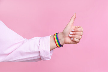 Woman wearing wristband in LGBT colors and showing thumbs up on pink background, closeup