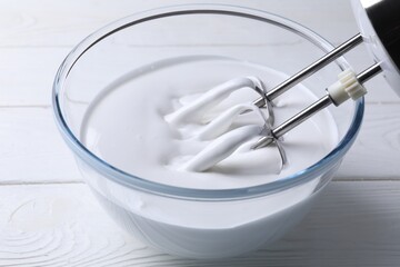 Mixing whipped cream with hand mixer in bowl on white wooden table, closeup