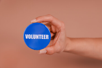Woman showing blue button badge with word Volunteer on light brown background, closeup