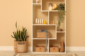 Fototapeta premium Shelving unit with dishware, jars of different products and other stuff near beige wall indoors
