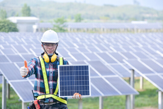 Engineer holding a solar panel standing in front of rows of solar panels at a solar powered station, Thailand