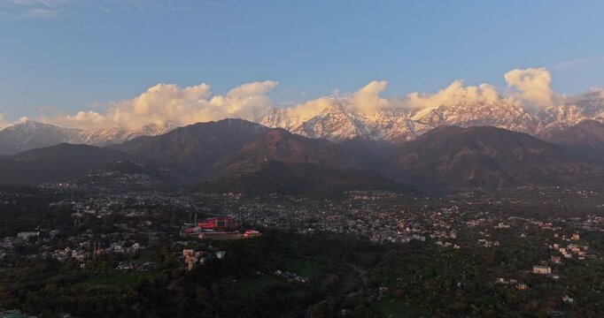 A beautiful aerial drone view of Dharamshala city, where the snow-covered Dhauladhar mountains gleam in the evening light, and the stunning Dharamshala Cricket Stadium shines in the heart of the city.