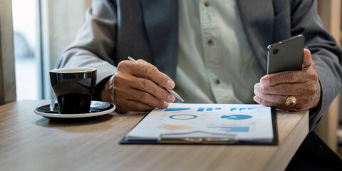 Senior Analysis and Communication. A senior man reviewing business charts while holding a smartphone in a cafe.
