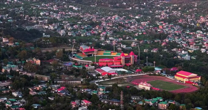 A left-to-right aerial panoramic view of Dharamshala city captures the Dharamshala Cricket Stadium and the athletics synthetic track glowing beautifully in the soft evening light.