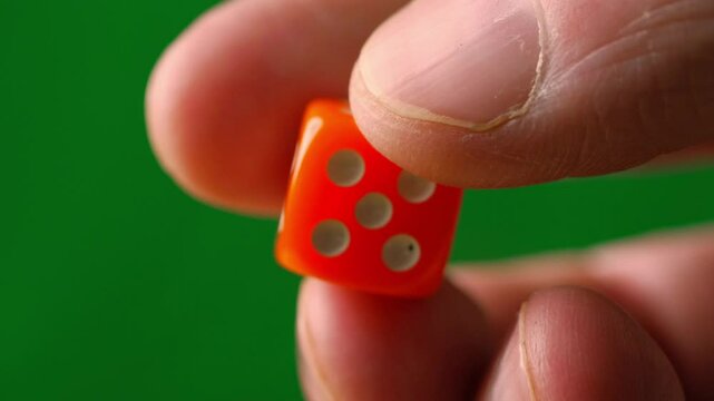 A close-up shot of a hand holding an orange die against a green background. The focus is on the die and the fingers holding it, showcasing a moment of chance or decision