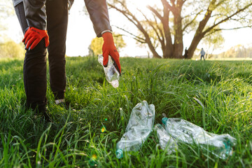 Volunteer wearing gloves collecting plastic bottles from green grass in park, promoting...