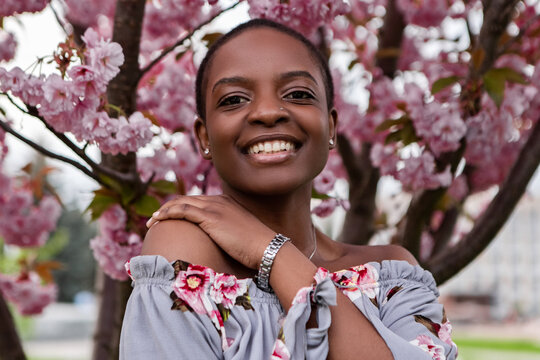 Happy young black woman with short hair and floral dress smiling under a blooming cherry blossom tree in spring, enjoying the beauty of nature. Japanese Sakura Flower